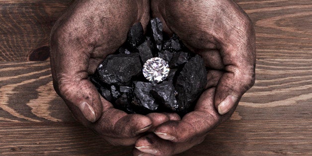 A miners hands holding a solitaire diamond in amongst pieces of coal.