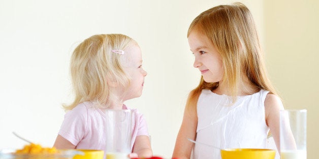 Two cute little sisters eating cereal with strawberries and drinking milk in white kitchen