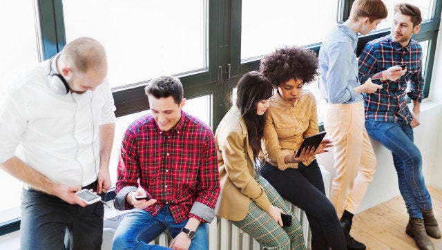 A group of young business people standing on a window talking and looking at their mobile devices. Backlit shot.