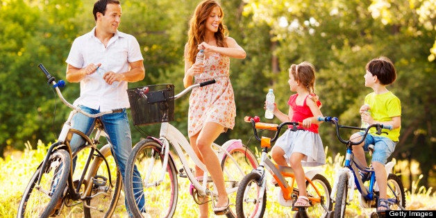 Young happy family riding bicycles outdoors in a park or nature. Making a break to refresh with water. Selective focus to little boy taking water bottle.