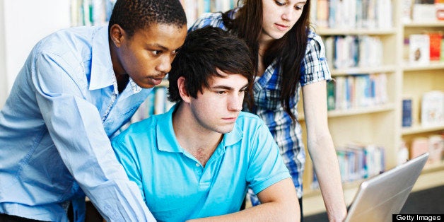 Three attractive young students, gathered around a library table with piles of books and a laptop computer they are sharing, concentrating on the image on the screen.