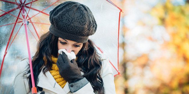Sad woman with cold or flu blowing her nose with a tissue under autumn rain. Brunette female sneezing and wearing warm clothes against cold weather. Illness, depression and allergy concept.