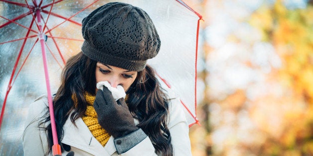 Sad woman with cold or flu blowing her nose with a tissue under autumn rain. Brunette female sneezing and wearing warm clothe