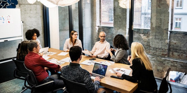 A group of young people, male and female, of different ethnicities are sitting in a bright modern office room. They are discussing something during a business meeting. There are documents and laptops on the table.