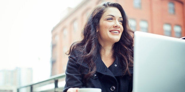 A happy young beautiful professional sits at an outdoor cafe in the city, holding a cup of espresso coffee and smiling while taking a break from her laptop computer. Horizontal with copy space. SELECTIVE FOCUS ON FACE.