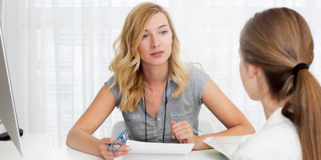 Blond haired businesswoman sitting at a desk opposite young woman and talking.