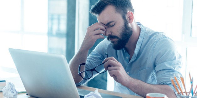 Frustrated young beard man massaging his nose and keeping eyes closed while sitting at his working place in office