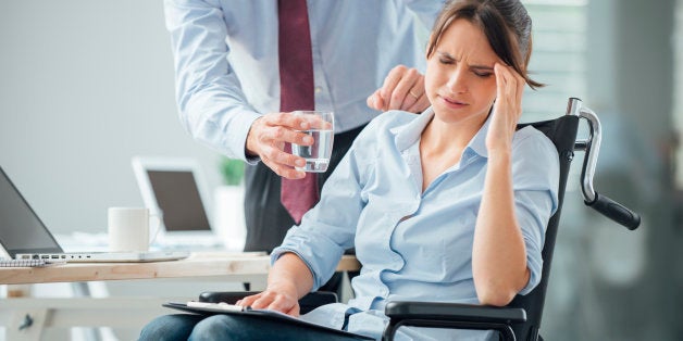 Business woman in wheelchair having an headache at office, her collegue is giving her a glass of water and helping her