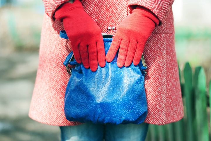 A fashion close up of a woman in a red spring-autumn wool topcoat, blue jeans and red gloves. Woman is holding a blue leather bag in her hands. On the background you can notice a green fence