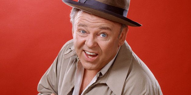 Carroll O'Connor (1924-2001), US actor, wearing a brown hat with a dark blue band and a beige jacket, and holding a cigar, in a studio portrait, against a red background, issued as publicity for the US television series, 'All in the Family', USA, circa 1975. The sitcom starred O'Connor as 'Archie Bunker'. (Photo by Silver Screen Collection/Getty Images)