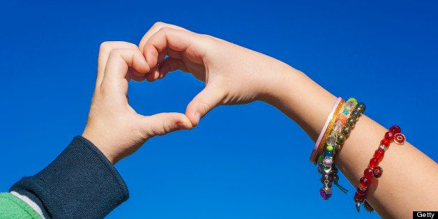 Young Girl With Bare Arms, Wearing Colorful Jewellery, And Young Boy Wearing A Shirt,Forming A Love Heart With Both Of Their Hands Together. Shot Against Blue Sky Background.