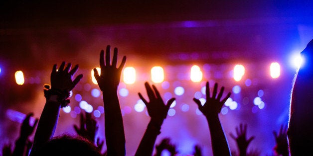 Audience with hands raised at a music festival and lights streaming down from above the stage. Soft focus, blurred movement.