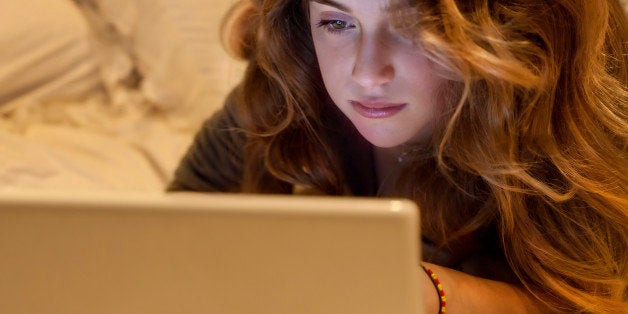 Beautiful young teenager on bed staring at screen of laptop and chatting on social networks with friends.