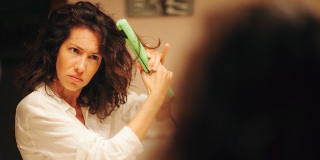 Disappointed woman using hair straightener in bathroom in front of mirror.