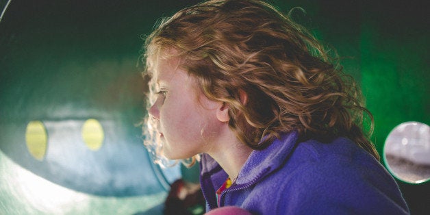 Little girl with curly hair sits in a play ground tube and looks a way from camera.