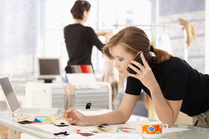 Young attractive female fashion designer leaning on office desk, talking on mobile phone.?