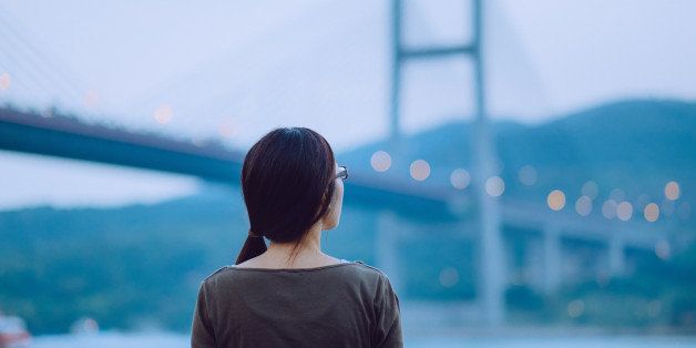 A rear view of a young woman looking away to the sea during sunset, with illuminated Tsing Ma Bridge in the background.