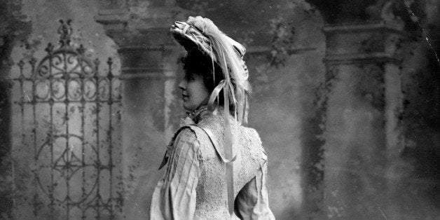 Miss Mansfield wearing a full sleeved coatdress, with lace detail, and a feather trimmed hat.