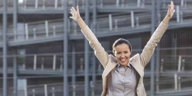 Excited young businesswoman with arms raised standing against office building