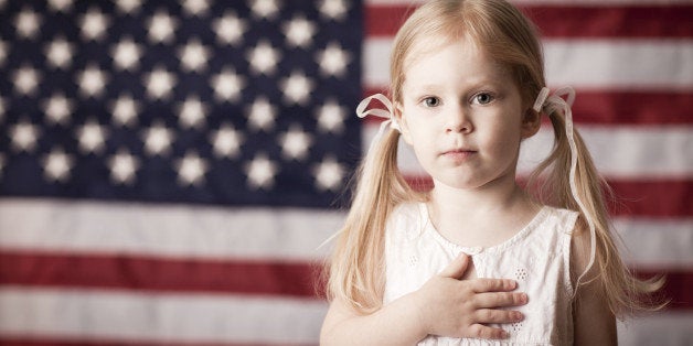 Color photo of a little blond-haired girl with her hand on her heart in front of an American flag.