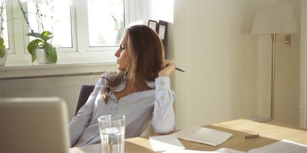 Beautiful caucasian female sitting at table with glass of water, documents and laptop. Business woman in home office looking away thinking.