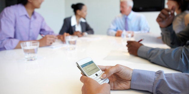 Woman using a Blackberry style mobile communication device to send a text message during a business meeting in a modern offic