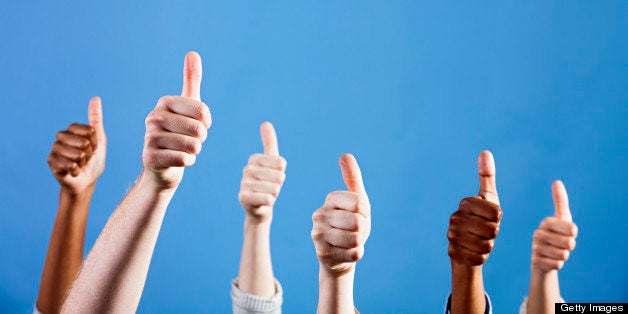 A group of six male and female hands of many different ethnicities join in giving a hearty thumbs up of approval against a plain blue background, with plenty of copy space. 