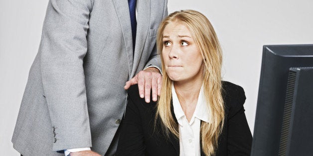 A businessman stands behind a pretty and vulnerable young businesswoman, leaning over her with one hand on her shoulder as she sits at her desk looking threatened, terrified and helpless.