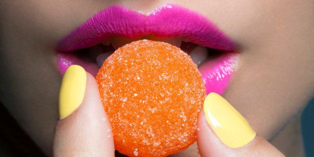 Young woman with pink lips and yellow nail polish holding an orange gumdrop in front of her lips, tight crop.