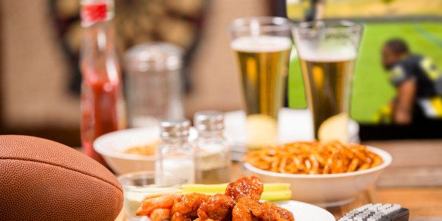 Hot wings and football in foreground. Beer in mugs in background with television. Football game on TV in a local pub or sports bar. Dartboard in background. Bar top. Superbowl party!
