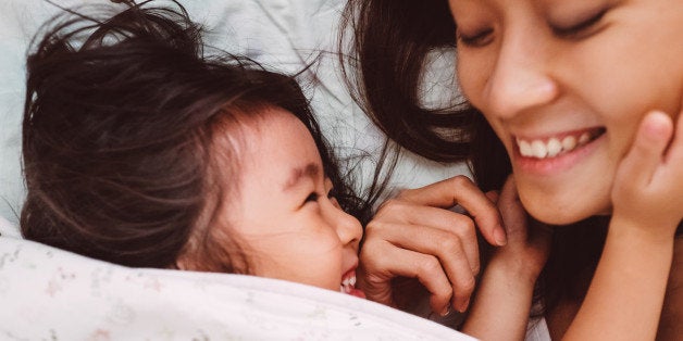 Lovely toddler girl putting her hands around her pretty young mom's face, while both looking at each other smiling joyfully on the bed after woke up in the morning.