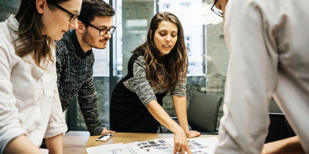 A team of young people in a modern office is discussing their project. The group consists of two men and two women, one of the is pointing at some papers while the others pay attention.