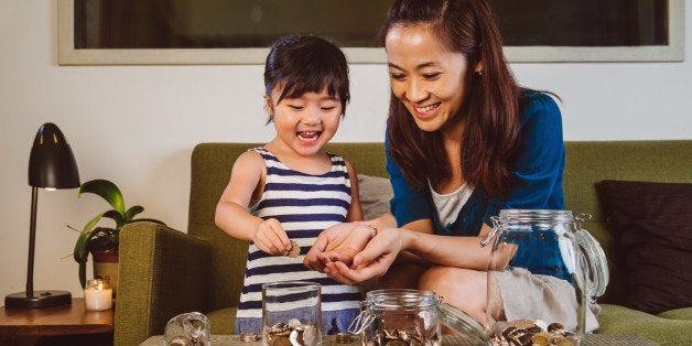 Lovely little girl putting coins into different glass jars on the coffee table joyfully under her pretty young mom's guidance at home.