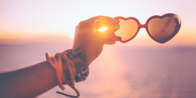 Cropped shot of a woman's hand holding a pair of heart-shaped sunglasses at sunset on a beach