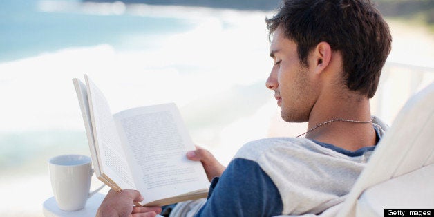 Man with coffee cup reading book in deck chair overlooking ocean