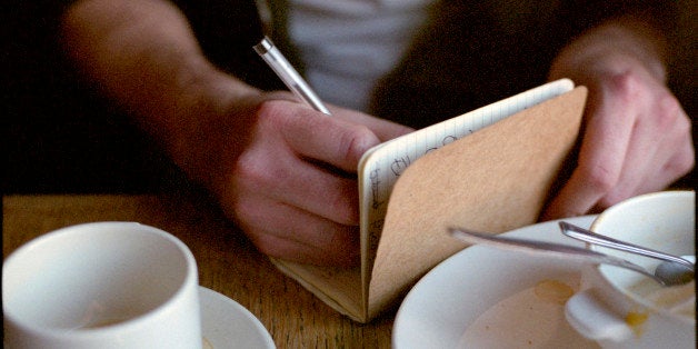 A writer sits in a cafe surrounded by teacups. He is writing in a notebook. The shot is a close up of his hands.