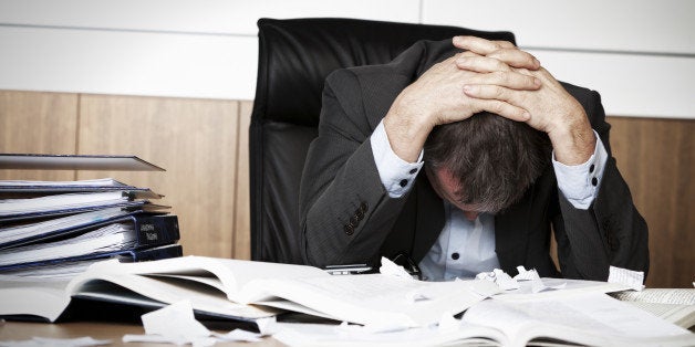 Worried businessman in dark suit sitting at office desk full with books and papers being overloaded with work.