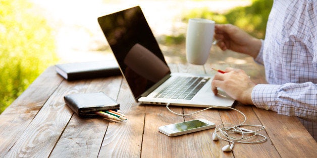 Casual dressed man sitting at wooden desk inside garden working on computer pointing with colour pen drinking coffee gadgets dropped around on table side view