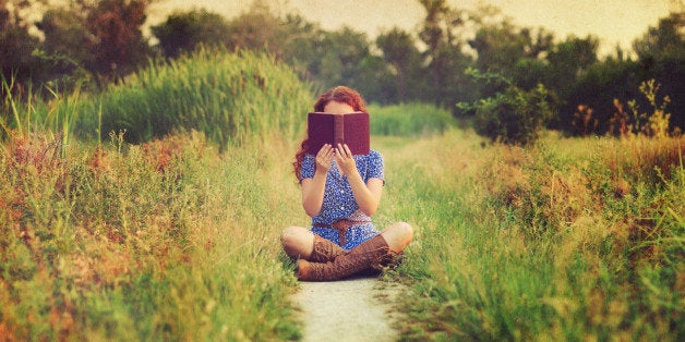 Young woman with long red hair wearing short-sleeved blue and white dress with brown belt and mid-calf brown leather boots sits cross-legged on narrow dirt footpath through field and holds up old brown book with both hands, obscuring face. The background is in shallow focus and overlaid with old paper texture.