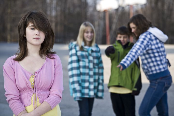 Alone in school, young girl standing away from schoolyard friends as they make fun of her behind her back