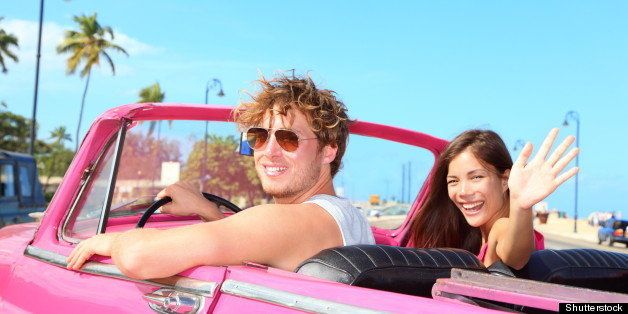 Couple happy in vintage retro convertible car. Friends driving on summer road trip in pink car. Beautiful young Caucasian man and Asian woman smiling joyful on sunny summer day traveling.