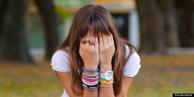 Young girl in fashionable warn out jeans, snickers and t-shirt hides her face with hands while sitting in the park on nice autumn day.