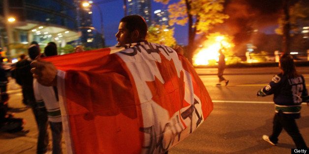 VANCOUVER, BC - JUNE 15: A person with a Candanian flag walks in front of a burning vehicle on June 15, 2011 in Vancouver, Canada. Vancouver broke out in riots after their hockey team the Vancouver Canucks lost in Game Seven of the Stanley Cup Finals. (Photo by Elsa/Getty Images)