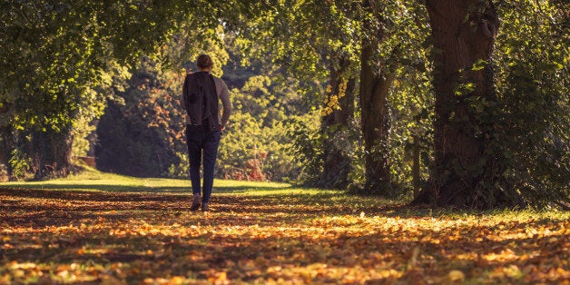 Solitary woman wanders contemplatively through a autumnal tree lined pathway with her jacket slung over her shoulder and the sunlight catching the fallen leaves scattered all around.