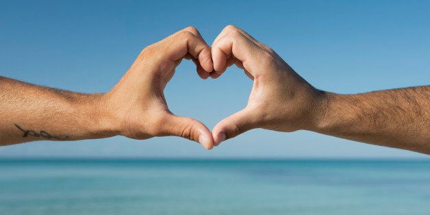 Two men's hands forming heart shape by ocean, cropped
