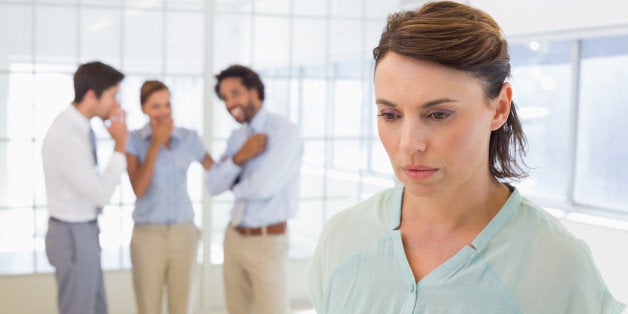 Colleauges gossiping with sad young businesswoman in foreground at a bright office