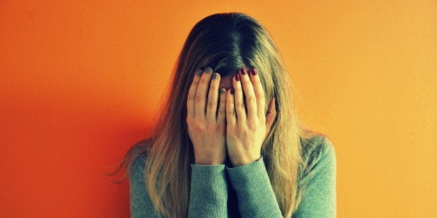 Woman closing her face with her hands, she has different red and blue nailpolish, standing against bright orange wall