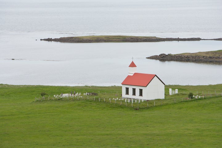 Small, white church at the edge of a body of water, surrounded by green grass, Iceland.