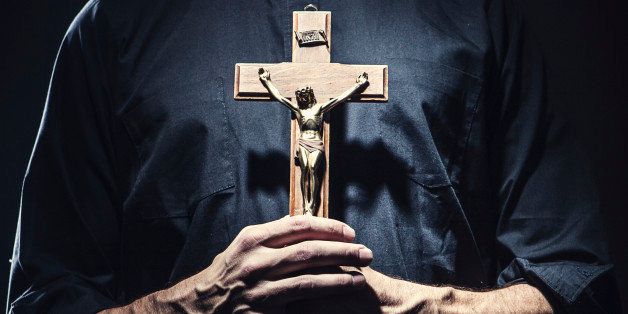 A dark high contrast image of a clergy man in a clerical collar holding a wooden portrayal of the crucifixion of Jesus Christ.