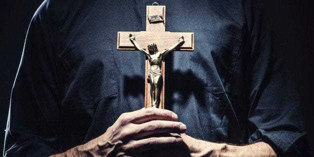 A dark high contrast image of a clergy man in a clerical collar holding a wooden portrayal of the crucifixion of Jesus Christ.